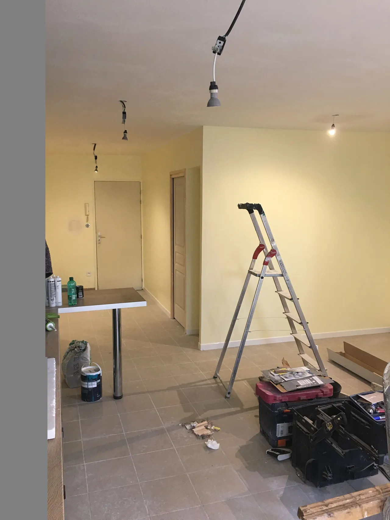 Séjour aux murs peints en jaune pâle, comptoir à pied chromé, porte d’entrée au fond, escabeau et caisses à outils au premier plan