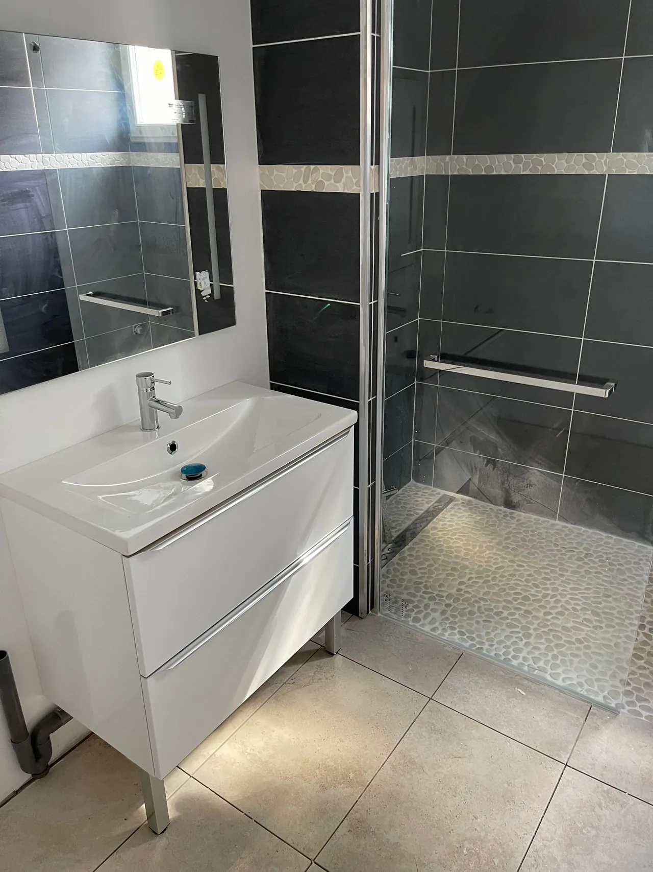 Salle d’eau terminée : meuble vasque blanc, miroir, douche à l’italienne au sol en galets, faïence anthracite