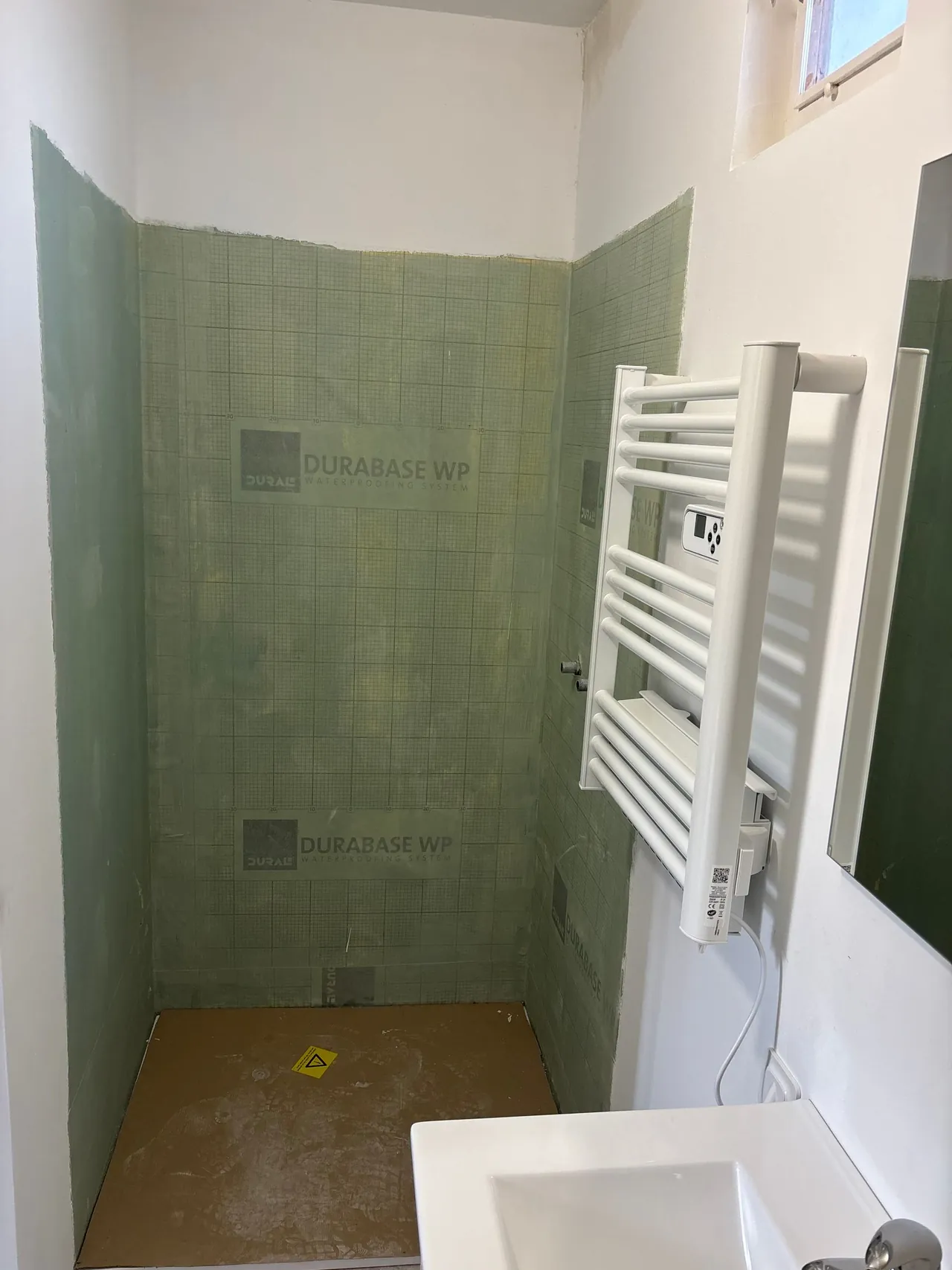 Salle d’eau en travaux : murs de la douche couverts de panneaux d’étanchéité verts, sèche-serviettes posé, vasque au premier plan