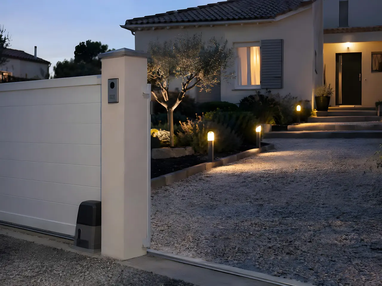 Portail coulissant blanc motorisé à la tombée du jour, visiophone sur le pilier et bornes lumineuses le long de l'allée