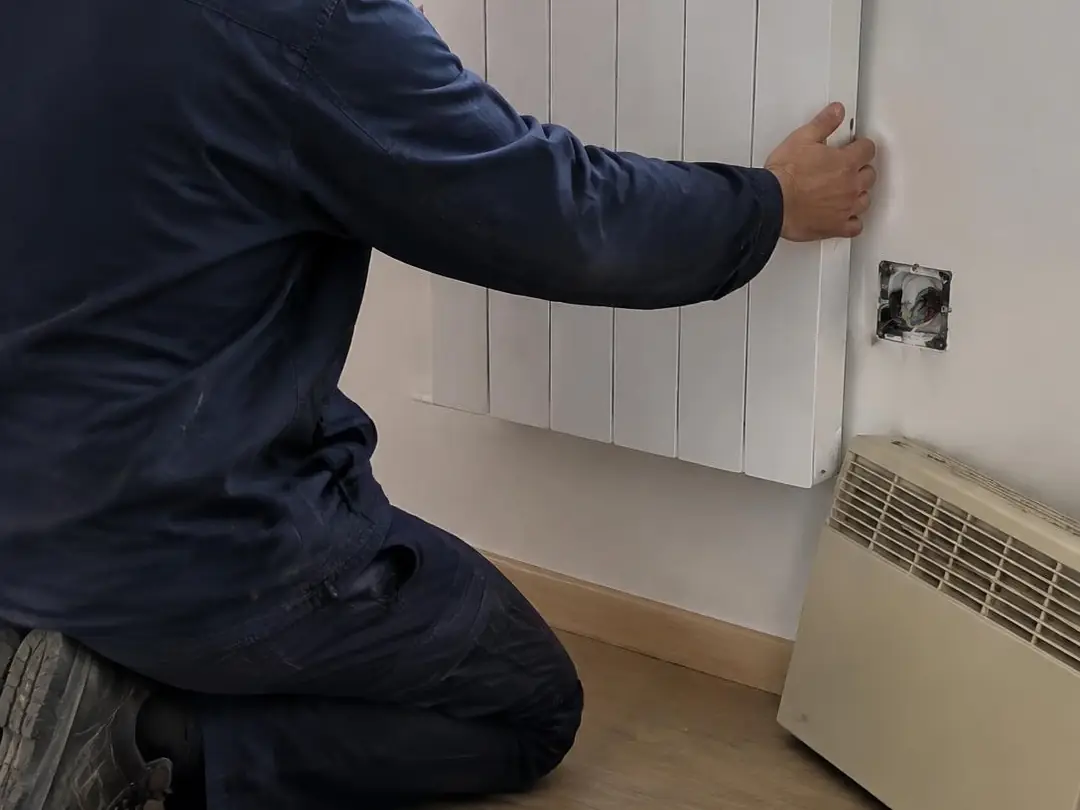Électricien à genoux qui fixe de niveau un radiateur neuf sous une fenêtre, vieux convecteur posé au sol