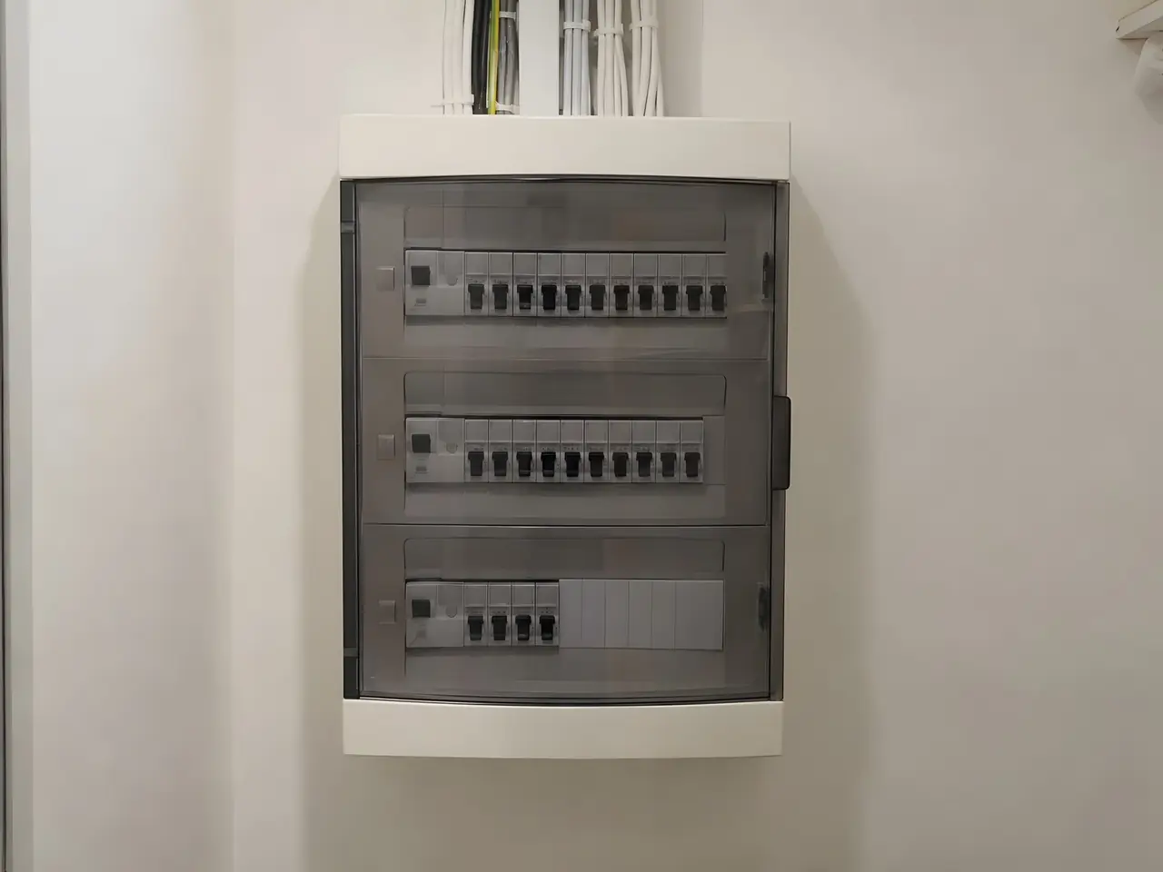 Tableau électrique blanc à trois rangées derrière une porte fumée, disjoncteurs alignés, places libres sous caches
