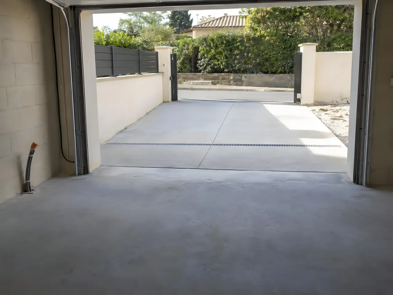 Intérieur d'un garage neuf vide : sol en béton, grande porte ouverte sur une allée bétonnée et un portail
