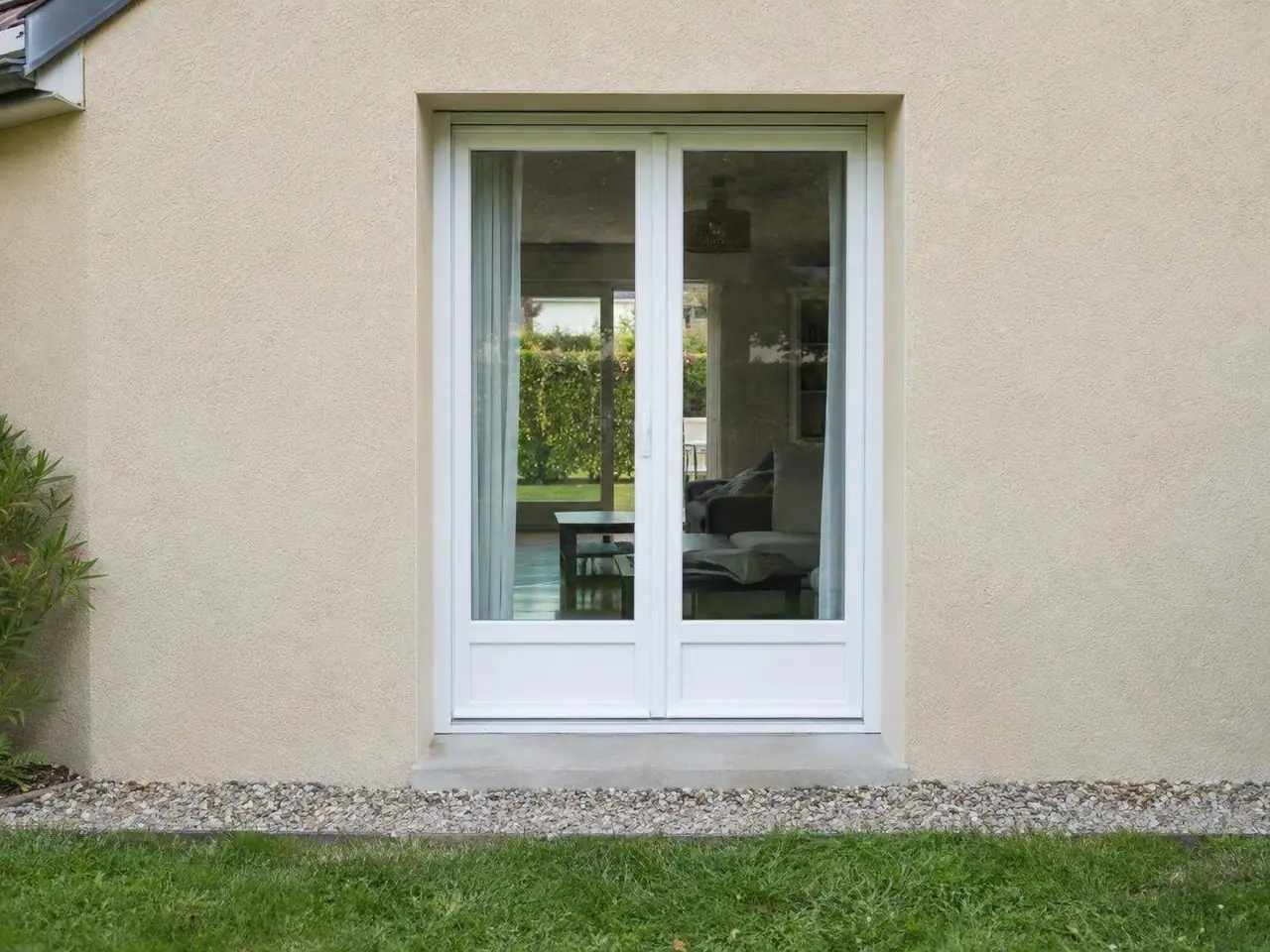 Porte-fenêtre blanche à deux vantaux sur un seuil en béton, façade en crépi beige, gravier et pelouse au pied