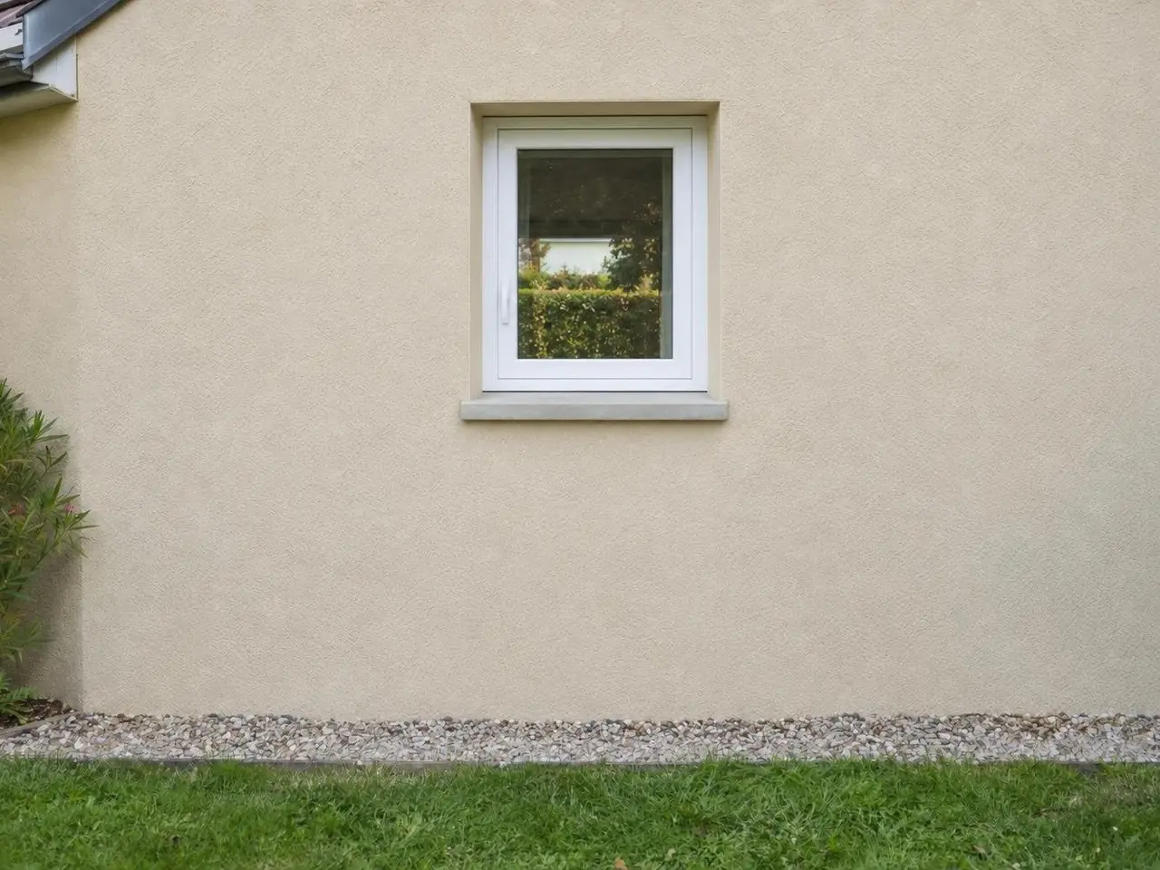Façade en crépi beige percée d'une petite fenêtre blanche à un vantail, bande de gravier et pelouse au pied