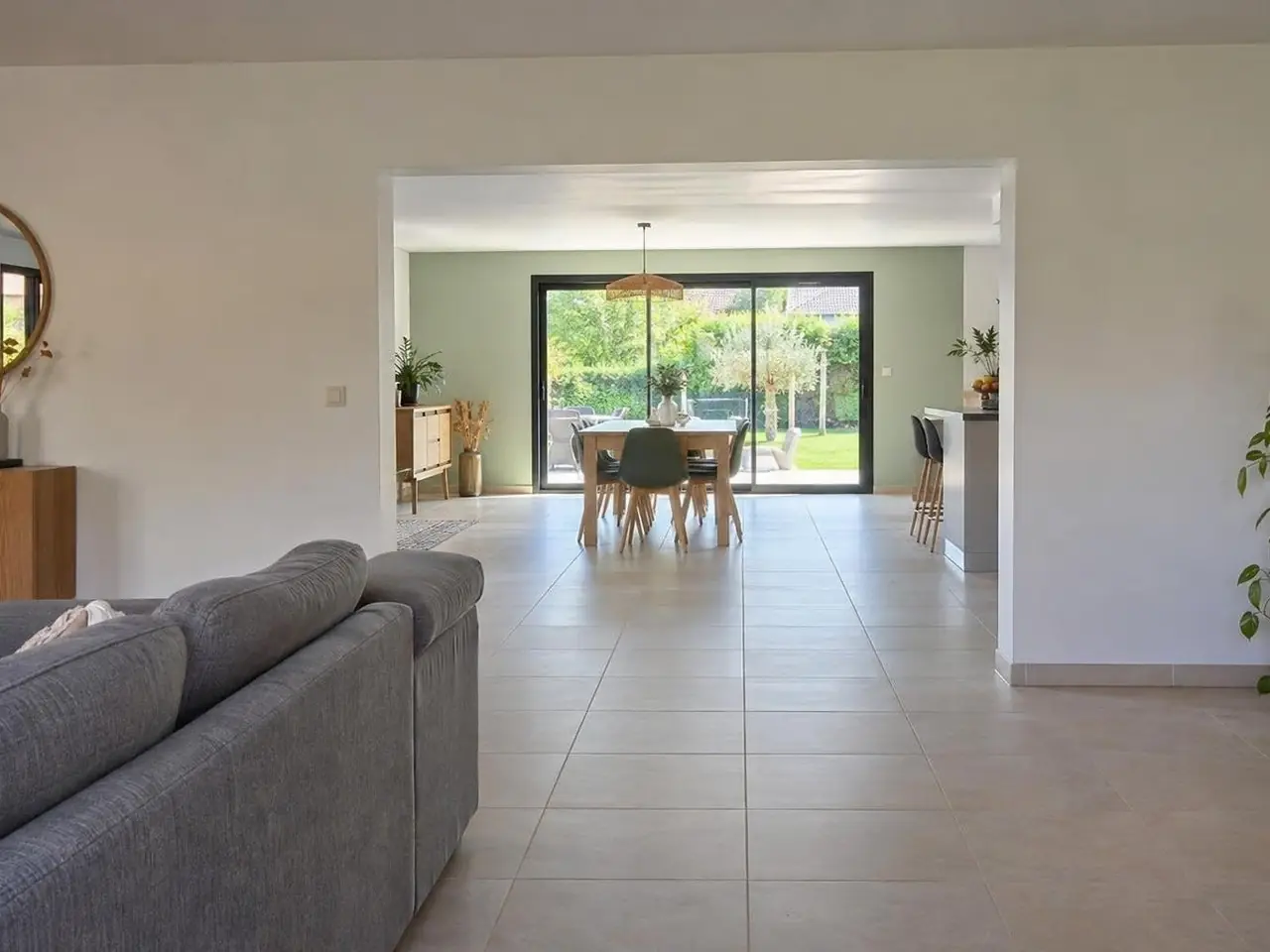 Séjour blanc au carrelage beige, canapé gris de dos, large passage ouvert sur une pièce neuve à baie vitrée