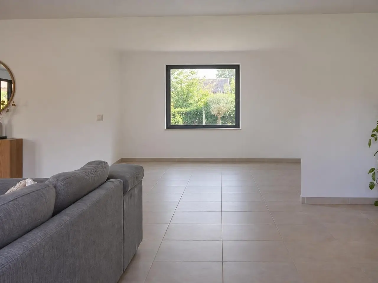Séjour blanc au carrelage beige, canapé gris de dos, mur du fond plein percé d'une fenêtre carrée sur le jardin