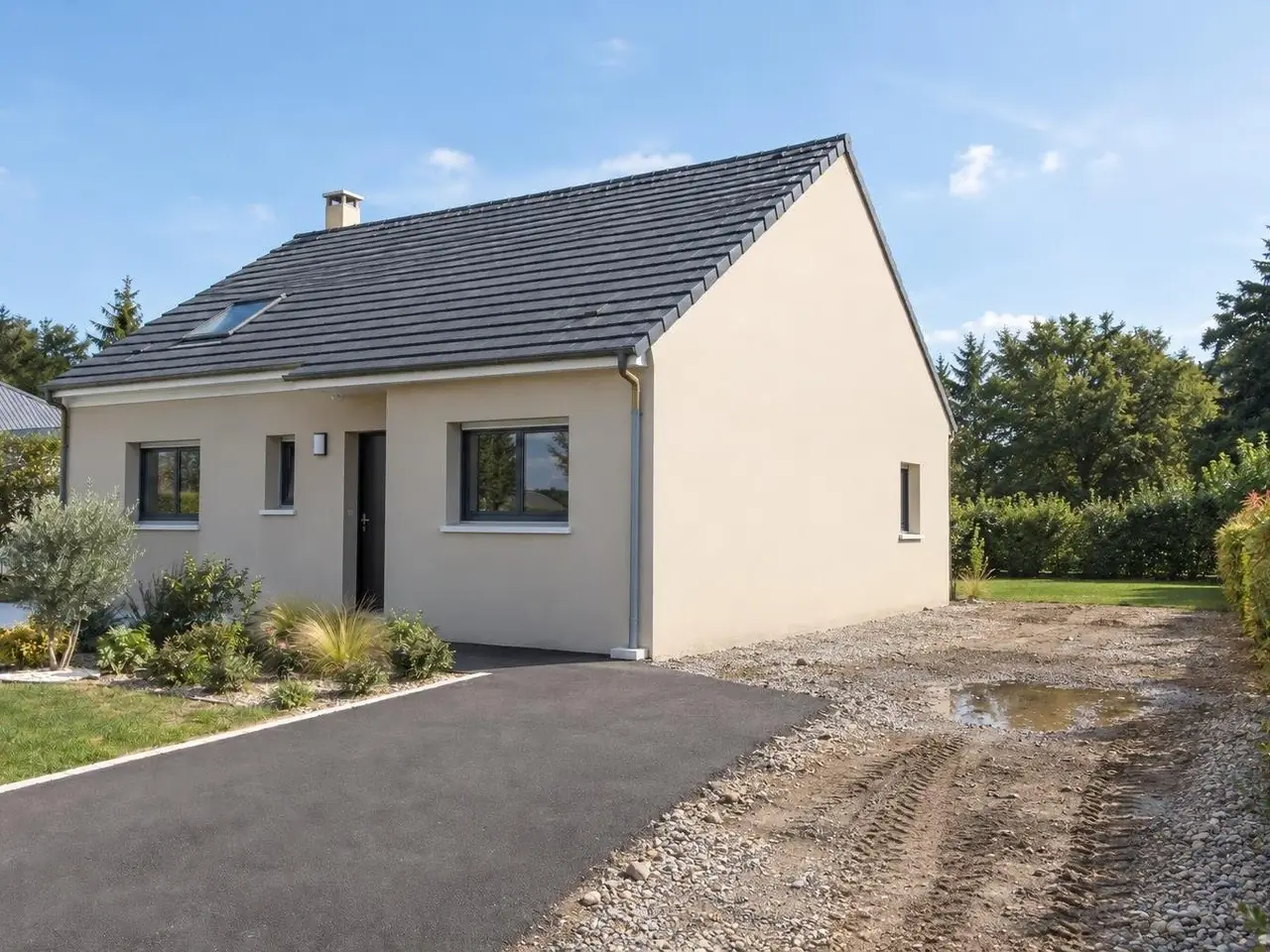 Pavillon crépi beige à toit de tuiles grises, pignon nu à petite fenêtre, terrain de gravier boueux et flaque à côté