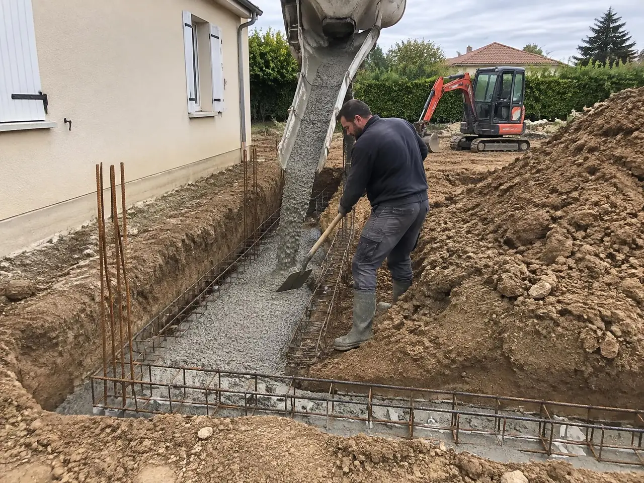 Toupie coulant le béton dans une fouille de fondation ferraillée le long d'un pavillon, maçon à la pelle