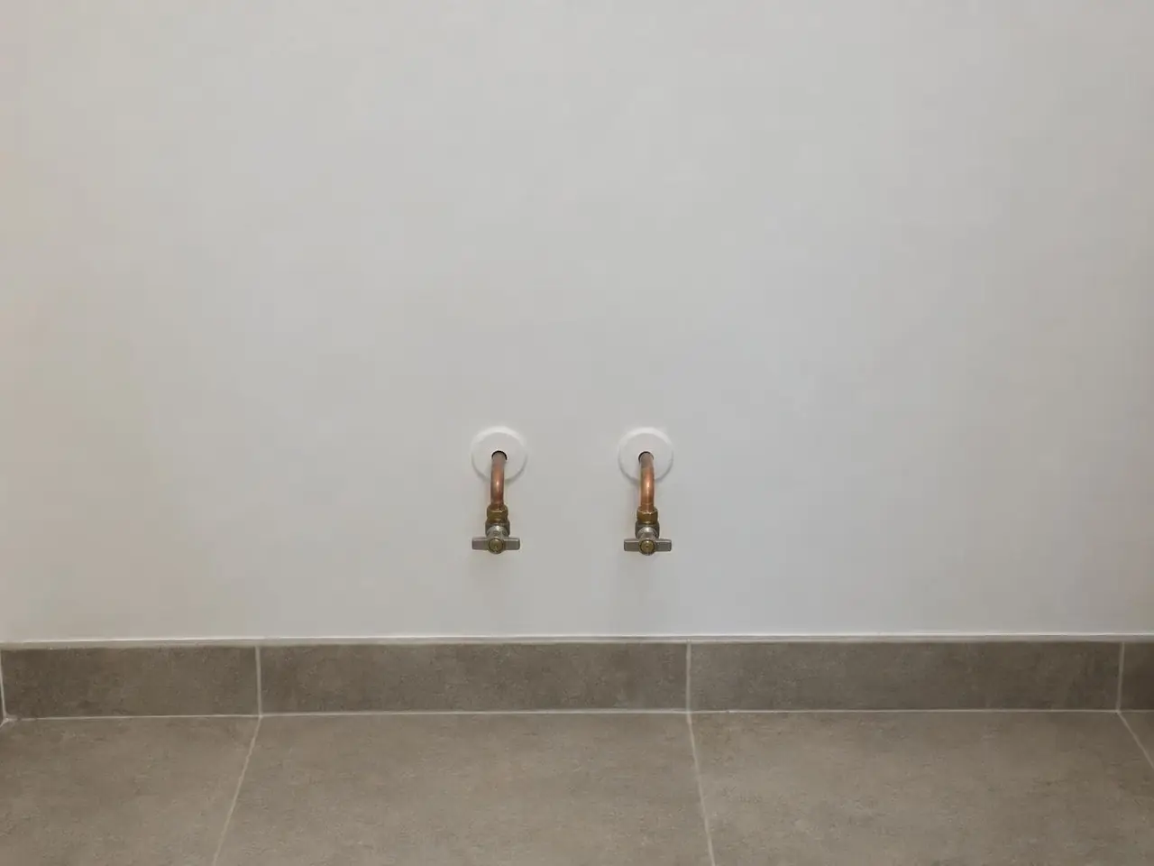 Mur blanc lisse de salle de bains, deux tuyaux en cuivre à robinets d'arrêt sortant de rosaces blanches, plinthe grise