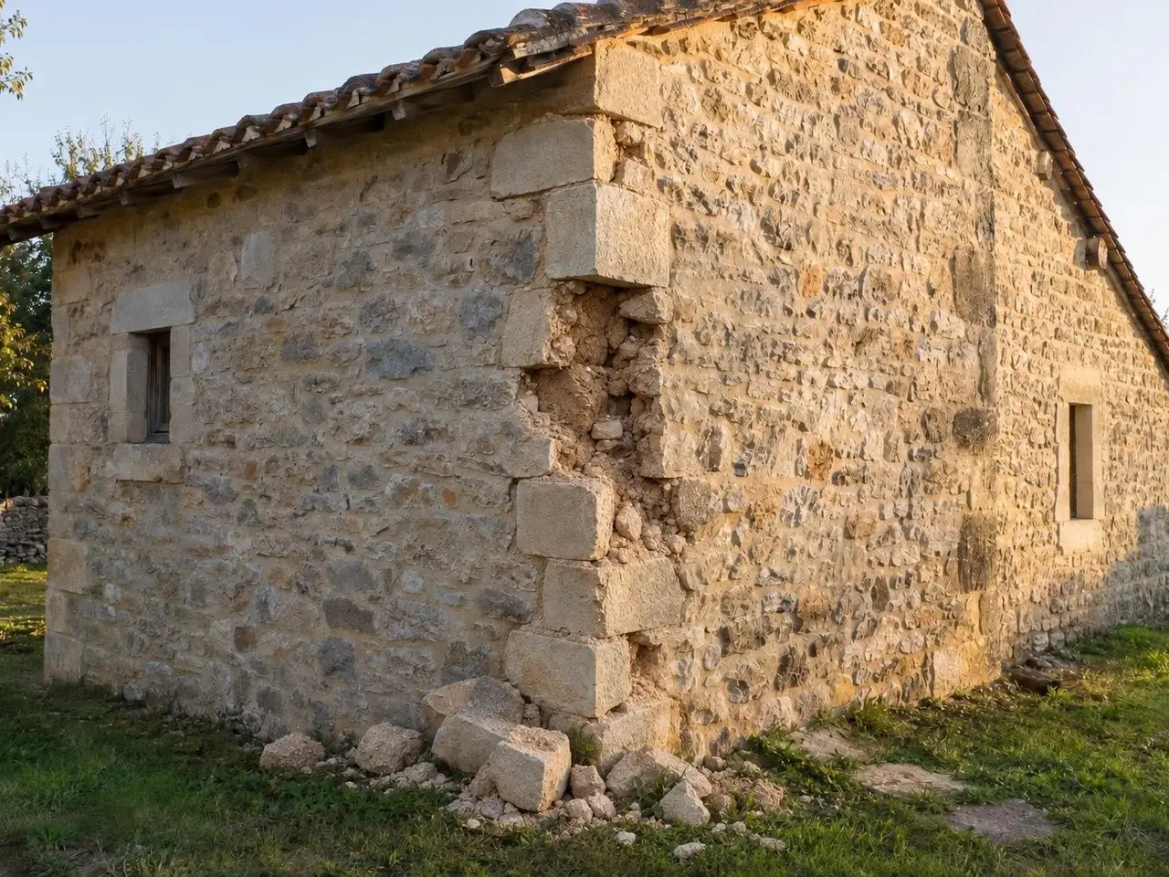 Angle d'une dépendance en pierre au toit de tuiles anciennes, pierres d'angle tombées dans l'herbe, blocage à nu
