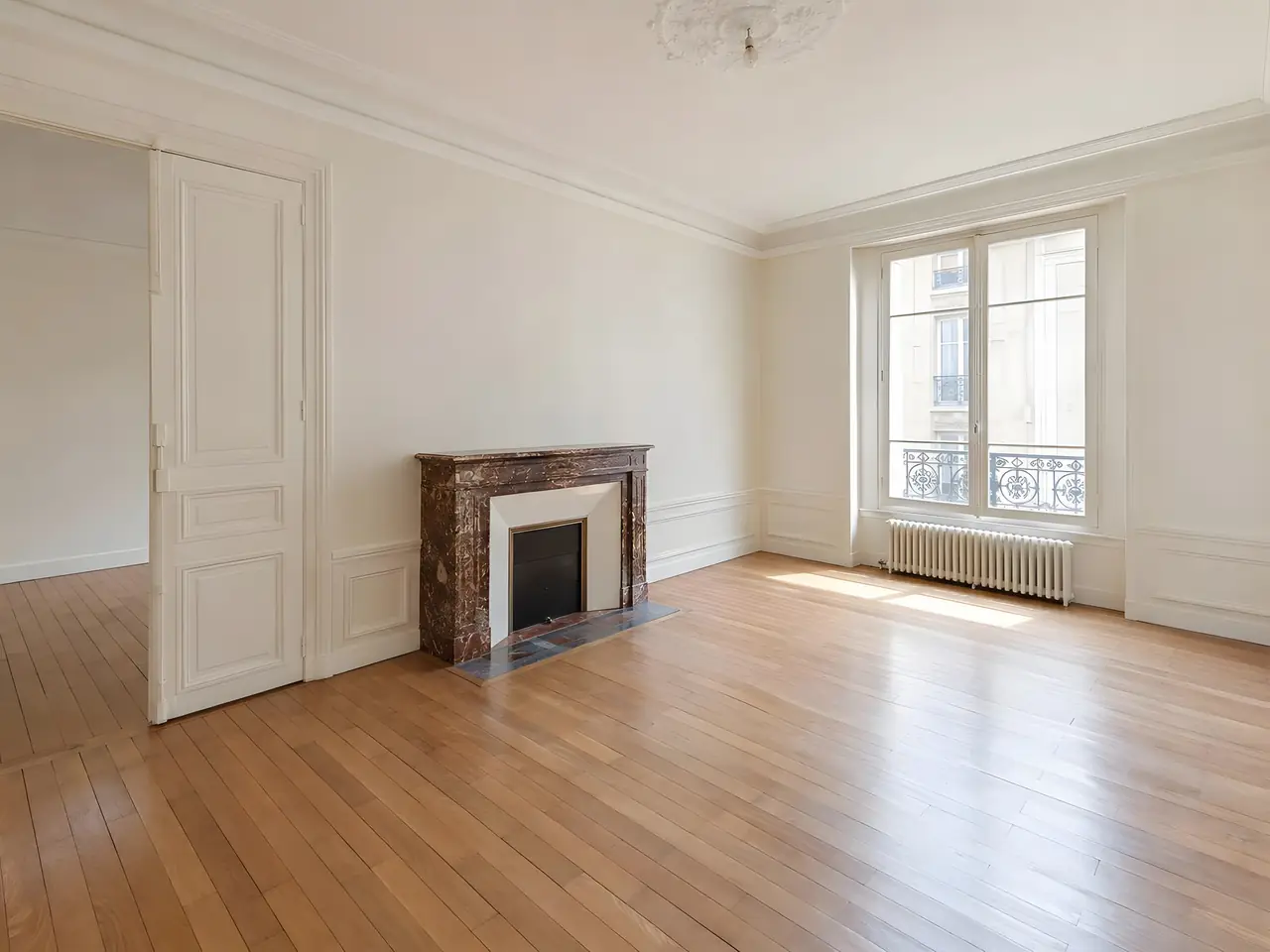 Grand séjour ancien et vide aux murs blancs et moulures, cheminée en marbre, parquet clair et porte-fenêtre à balcon