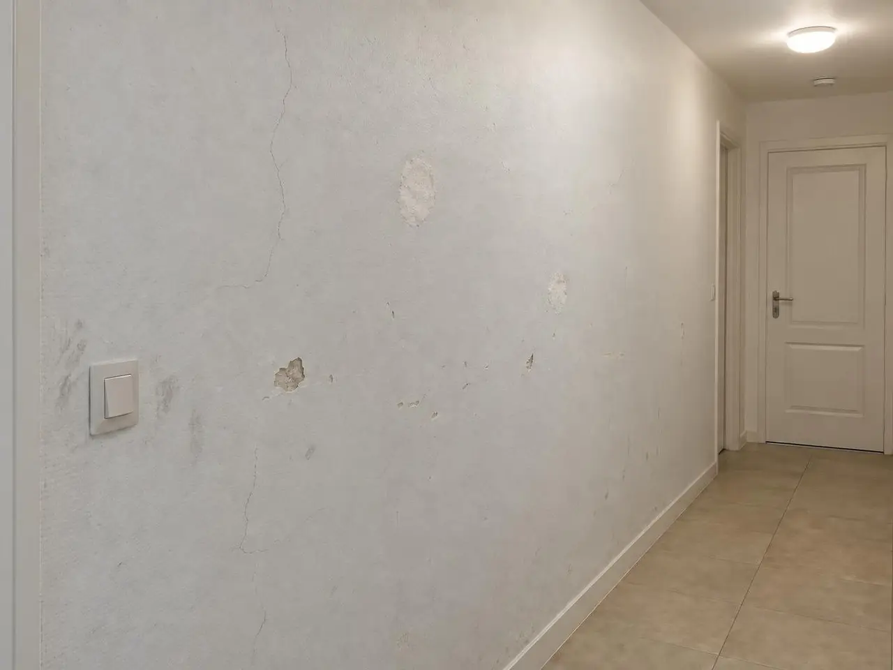 Mur de couloir peint en blanc grisé, fissures fines, éclats et rebouchages plus clairs autour de l'interrupteur