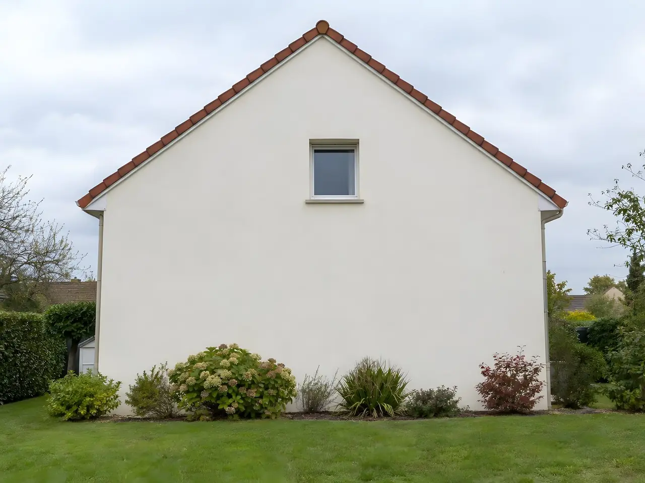 Pignon d'un pavillon repeint d'un blanc cassé uniforme sous un toit de tuiles rouges, massif d'arbustes au pied
