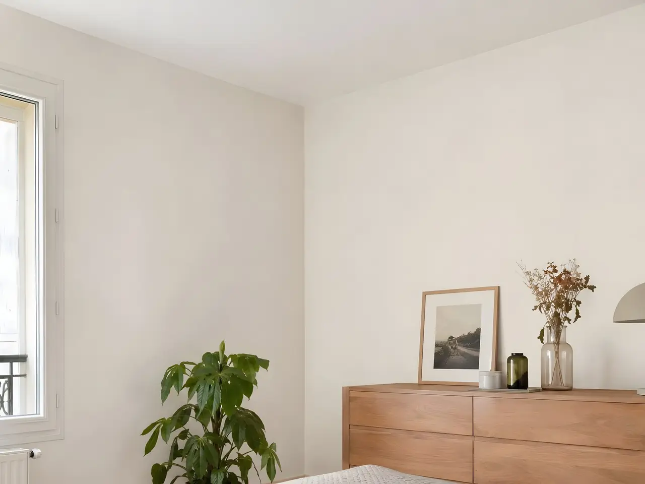 Angle de chambre aux murs blanc cassé sous un plafond blanc sans aucune trace, commode en bois et plante verte