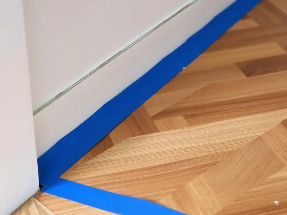 Adhésif de masquage bleu posé au ras d'une plinthe blanche, sur un parquet en bâtons rompus