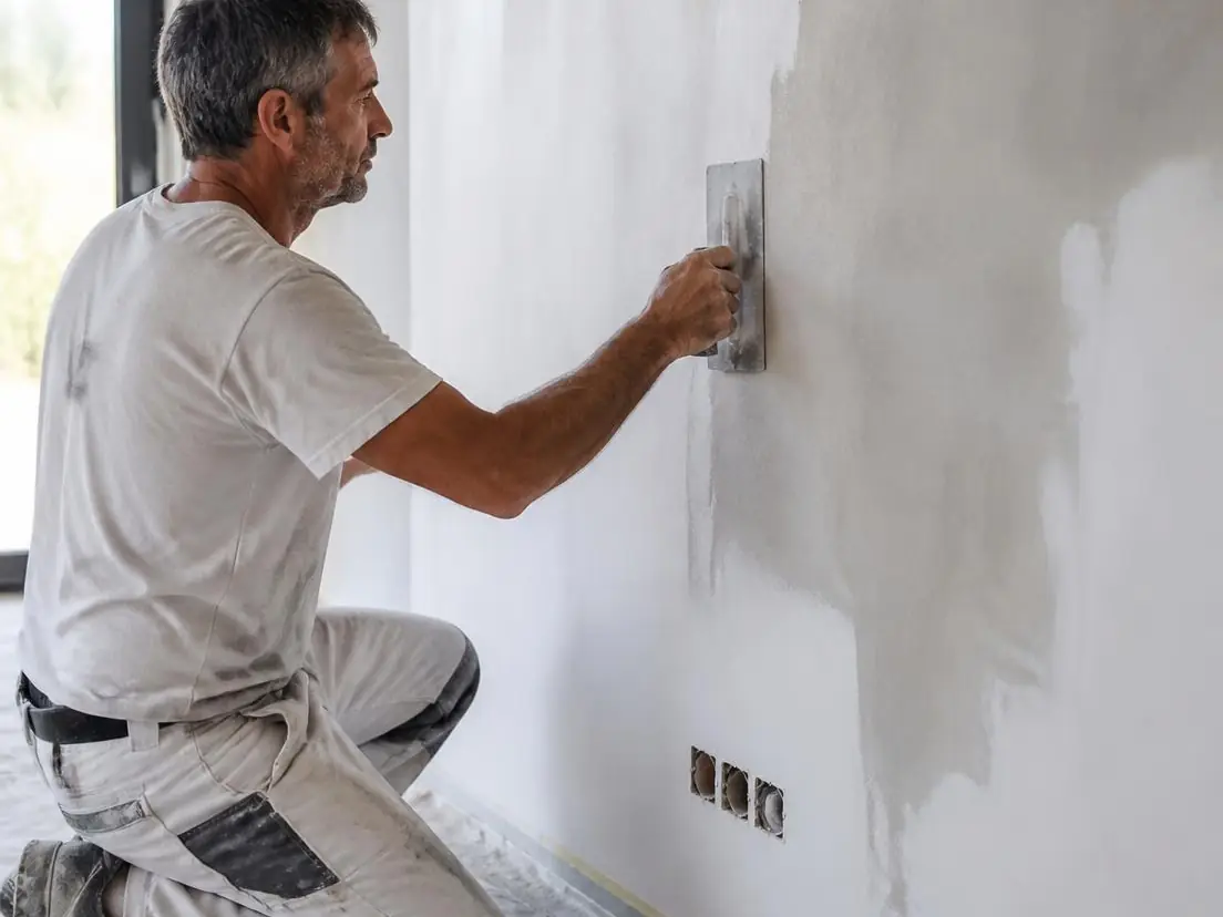 Peintre à genoux sur un sol bâché lissant l'enduit sur un mur blanc avec une lisseuse en inox