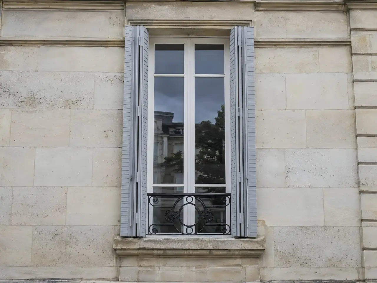 Persiennes métalliques repeintes en gris clair satiné, repliées dans le tableau d'une façade en pierre de taille