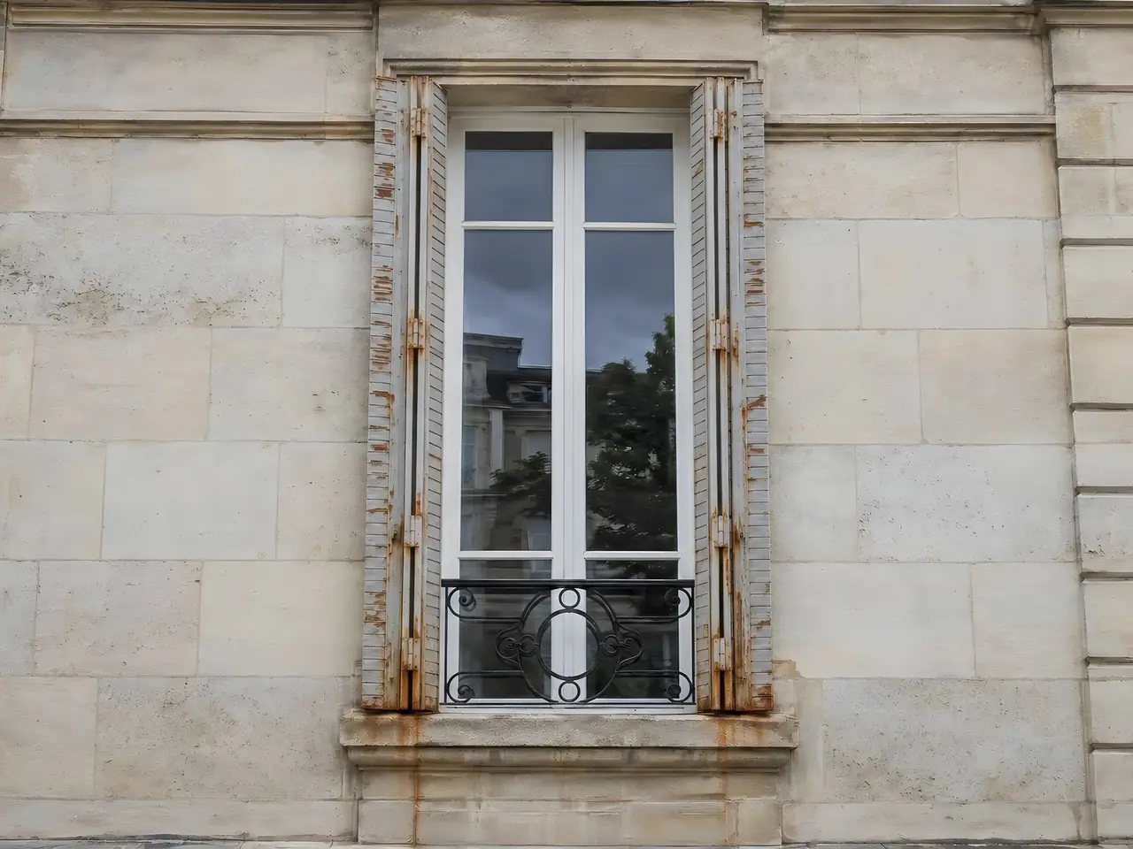 Persiennes métalliques grises écaillées et piquées de rouille, traînées orange sur la pierre sous la fenêtre