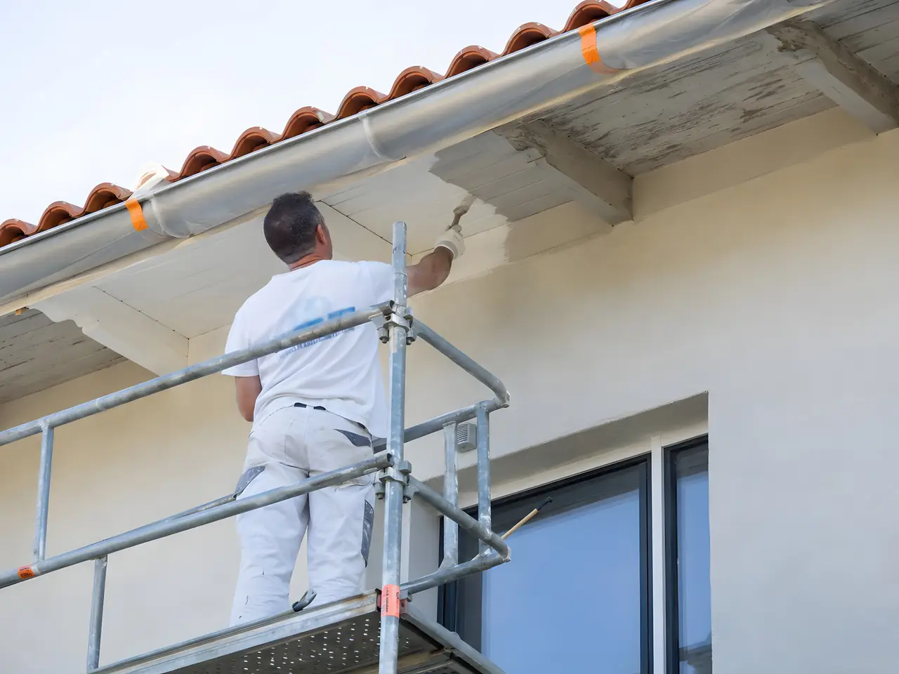 Peintre debout sur un échafaudage qui repeint en blanc au pinceau le dessous de toit d'une maison aux tuiles rondes