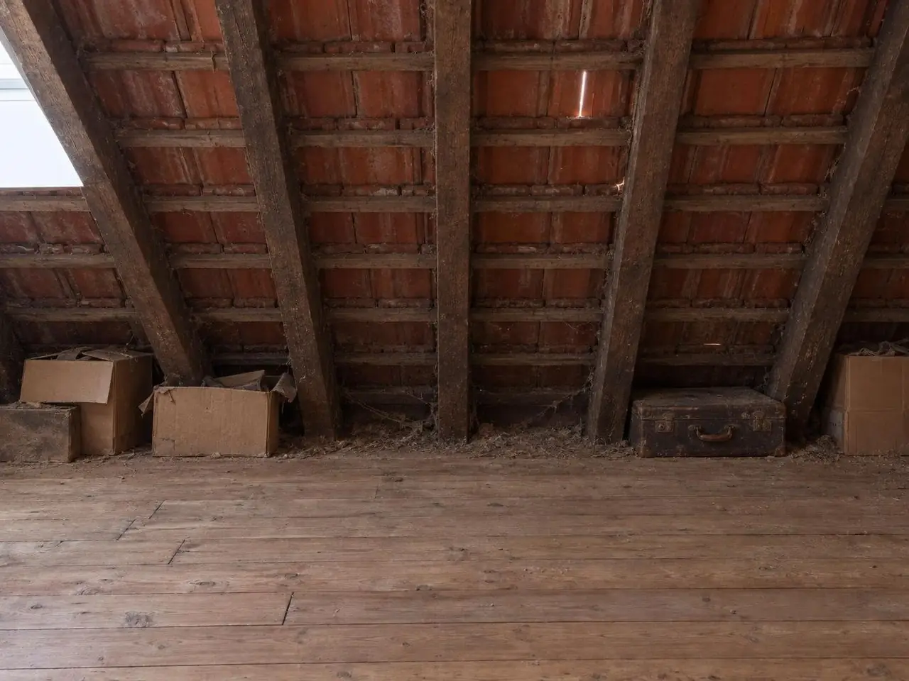 Pente de grenier aux chevrons sombres et dessous de tuiles apparents, cartons et vieille valise au pied du rampant