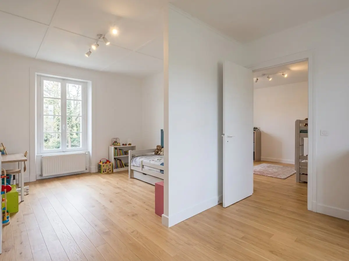 Cloison blanche qui partage une grande pièce en deux chambres d'enfant, porte ouverte de l'une à l'autre