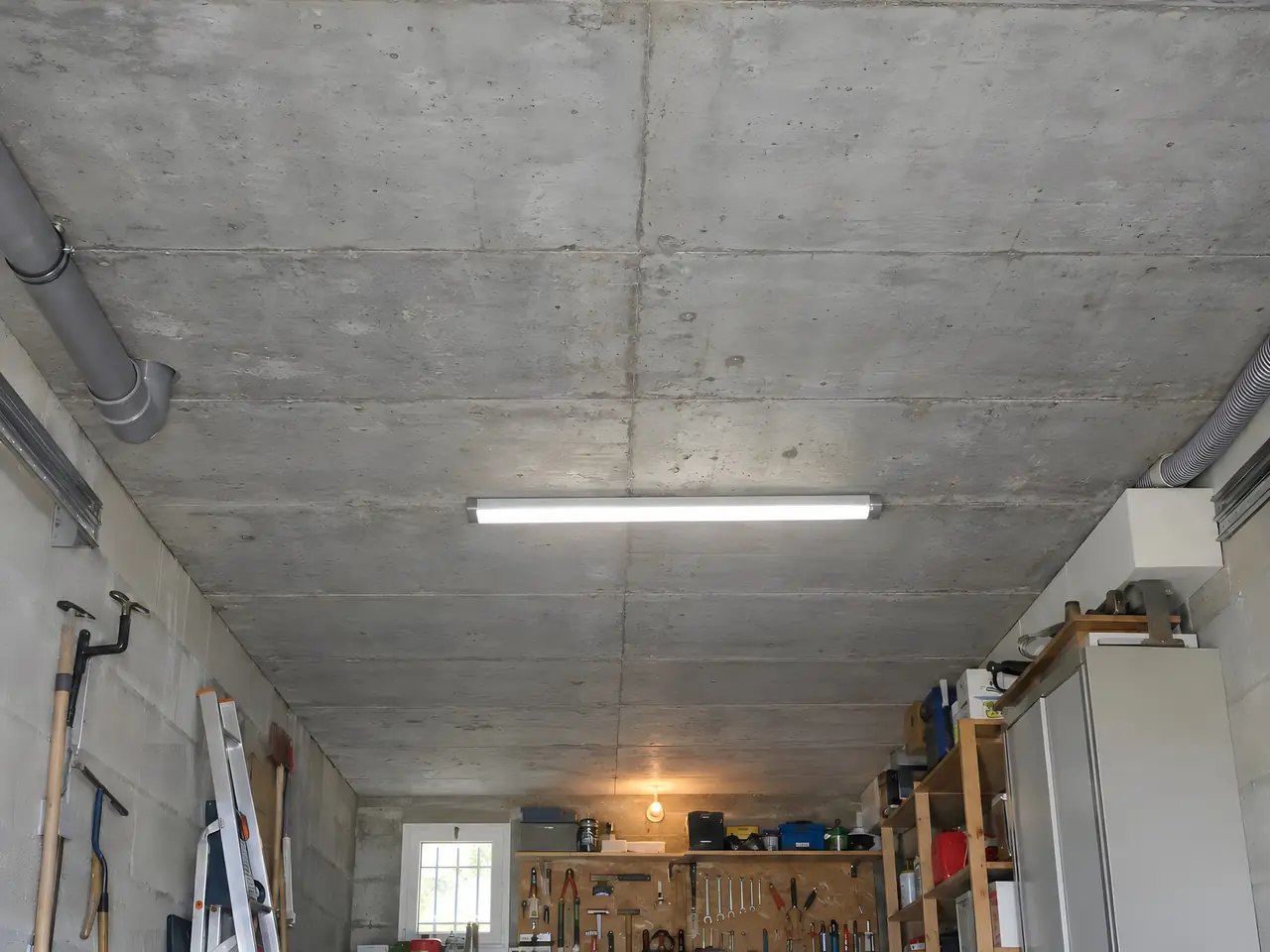 Plafond en béton brut d'un garage avec néon, gaines grises, échelle, outils au mur et établi au fond