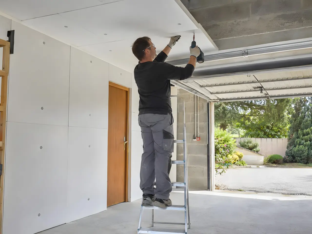 Artisan sur un escabeau qui visse des panneaux isolants blancs au plafond d'un garage, porte ouverte sur le jardin