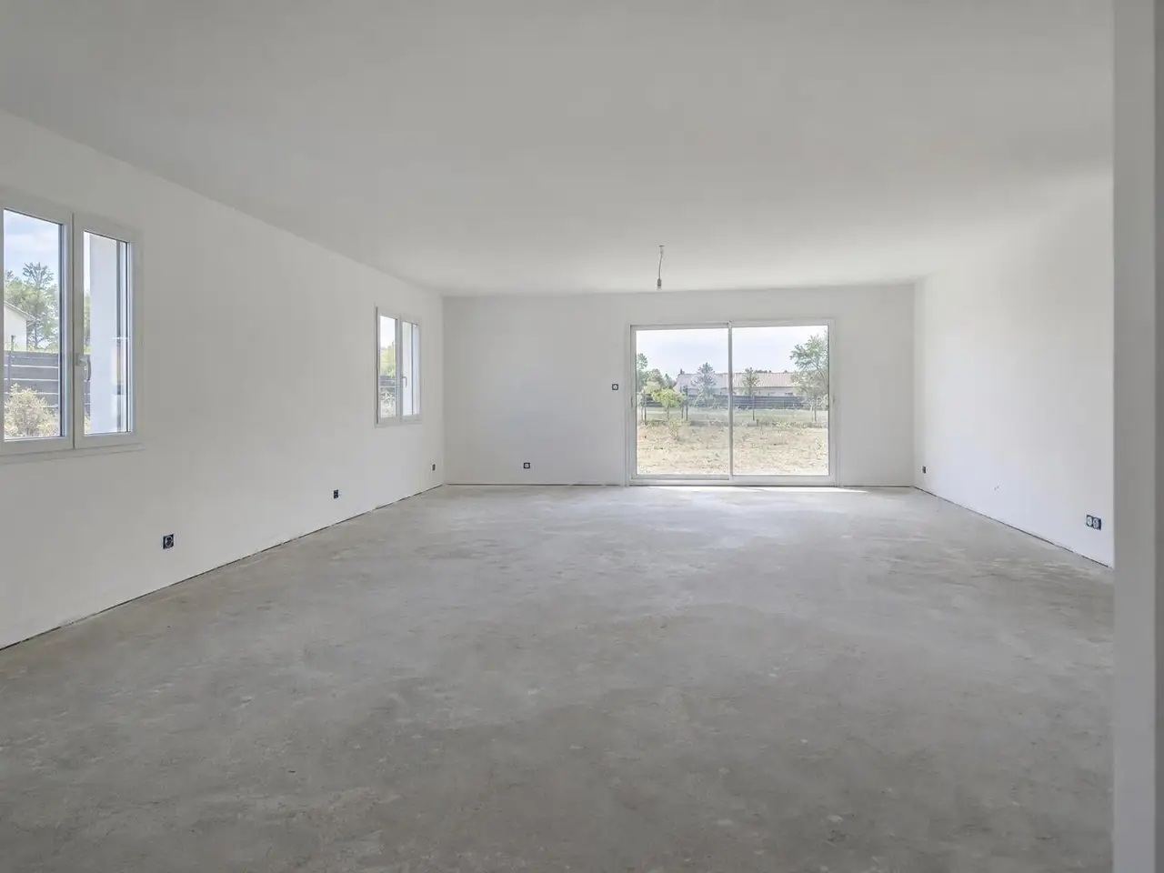 Séjour de maison neuve vide aux murs blancs et au plafond plat, chape de béton grise, baie vitrée au fond