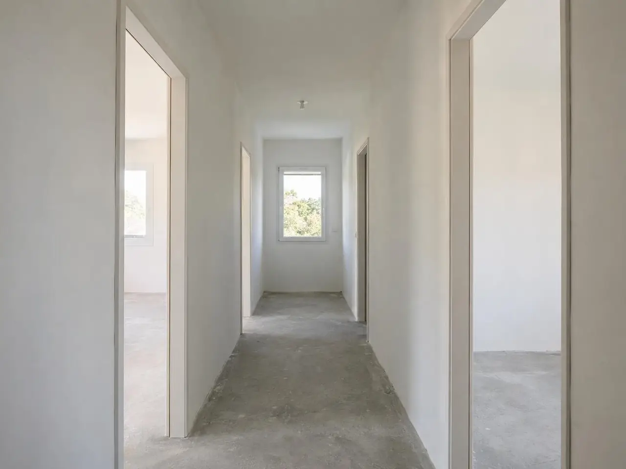Couloir aux cloisons blanches et lisses ouvert sur des chambres, passages de porte, fenêtre blanche au fond