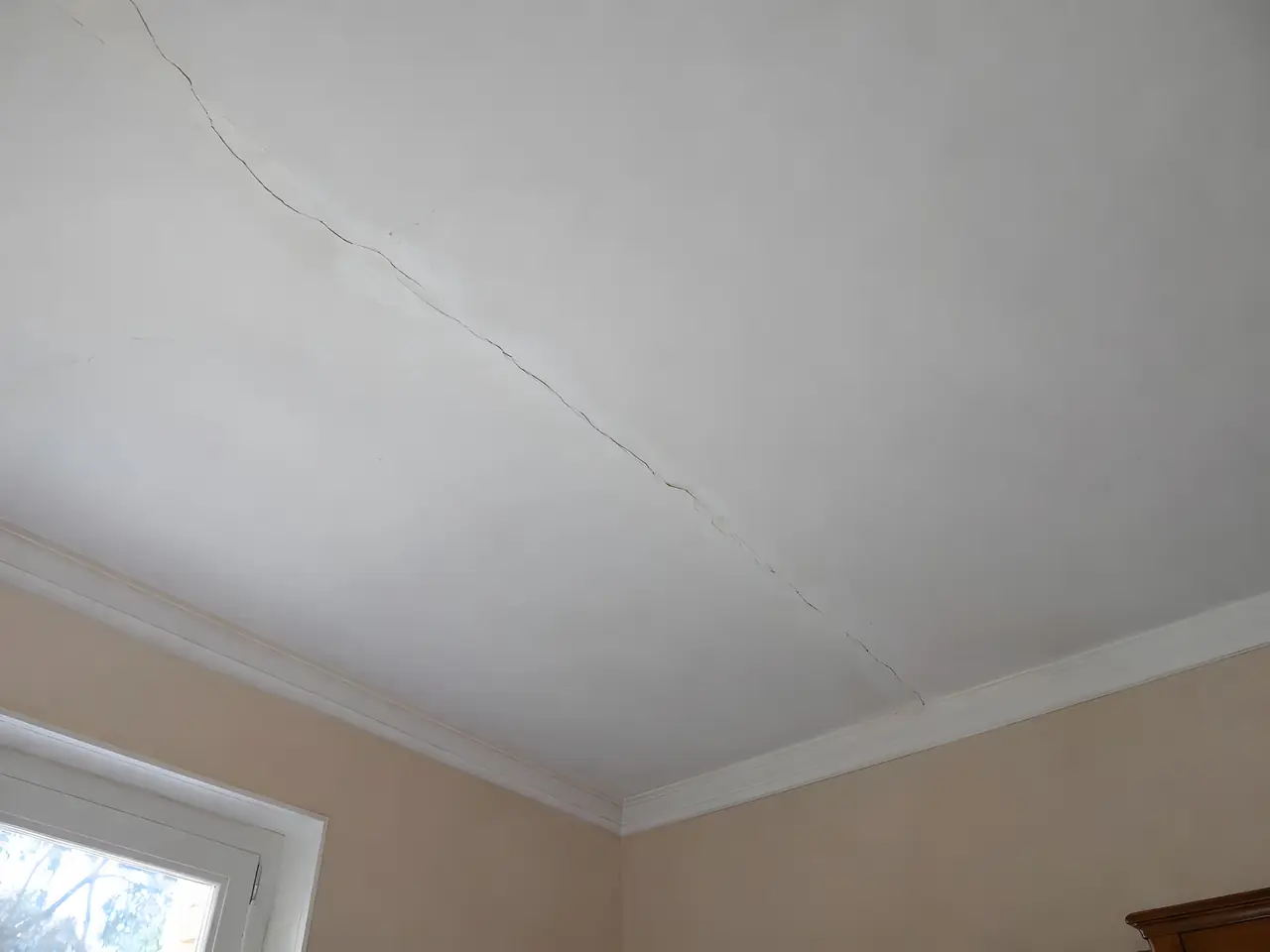 Plafond blanc ancien traversé d'une longue fissure en diagonale, au-dessus d'une corniche moulurée et de murs pêche