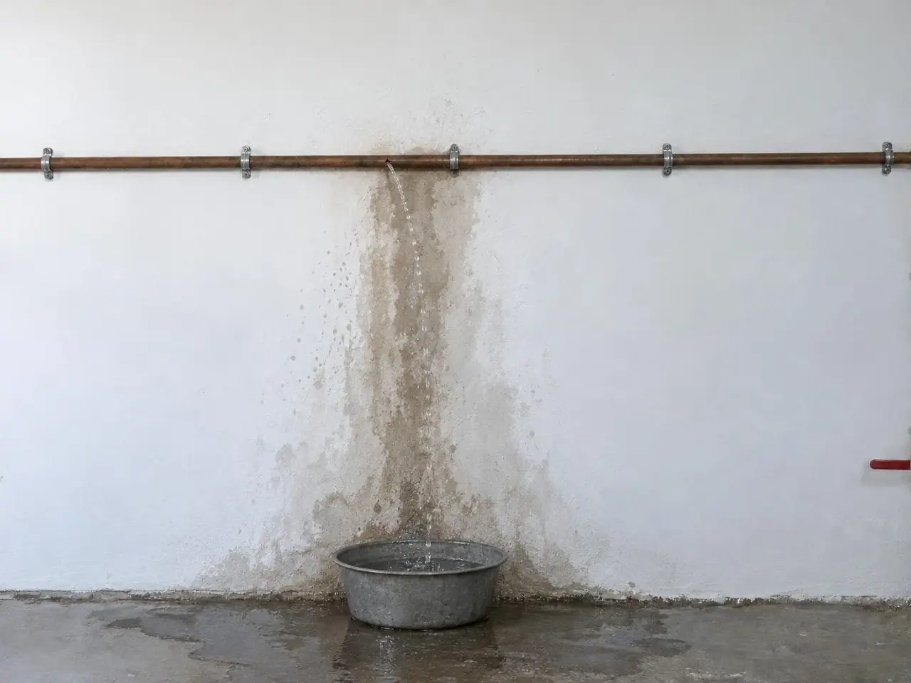 Tuyau de cuivre fendu qui projette un filet d'eau sur un mur blanc taché d'humidité, bassine et flaque au sol