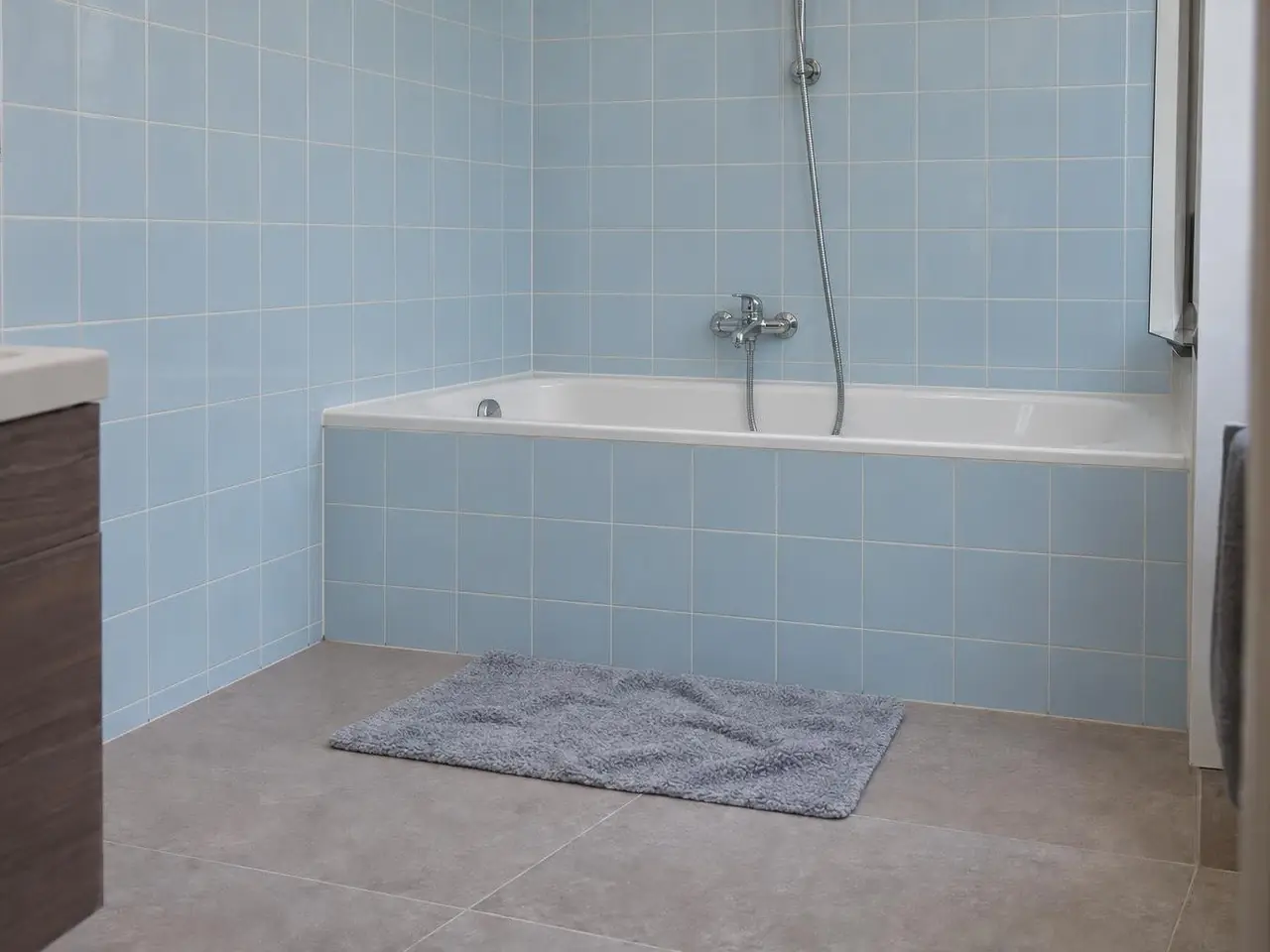 Salle de bains à faïence bleu pâle, baignoire encastrée dans un tablier carrelé, tapis de bain gris devant