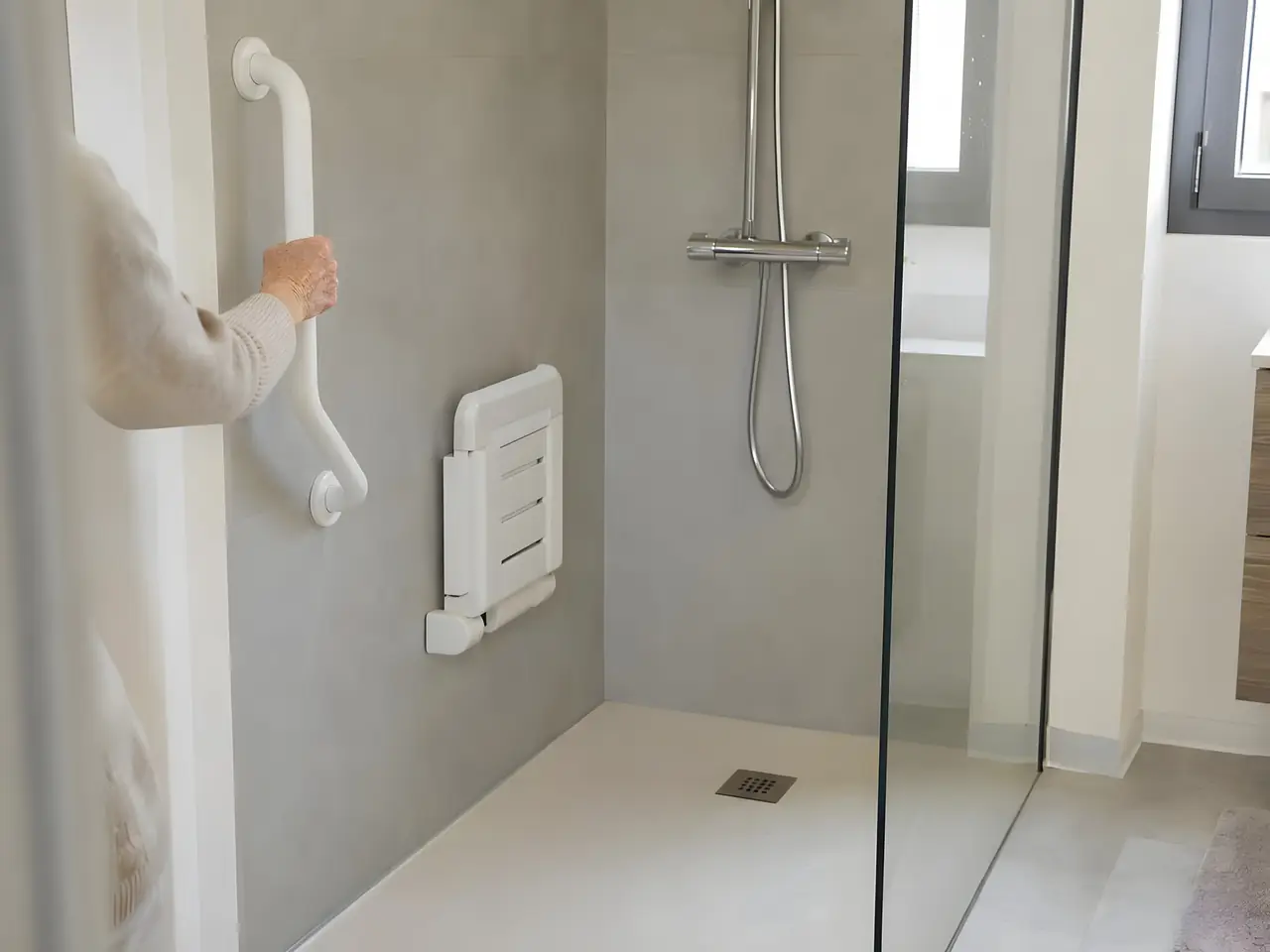 Douche de plain-pied avec barre d'appui blanche, siège rabattable et paroi vitrée, une main âgée tenant la barre