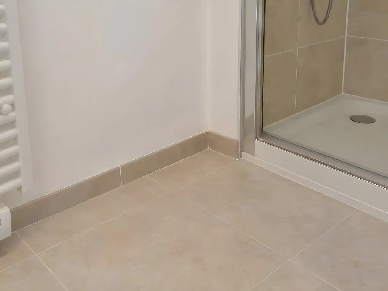 Coin de salle de bains au sol en carrelage beige refermé et intact, mur blanc propre à côté de la douche