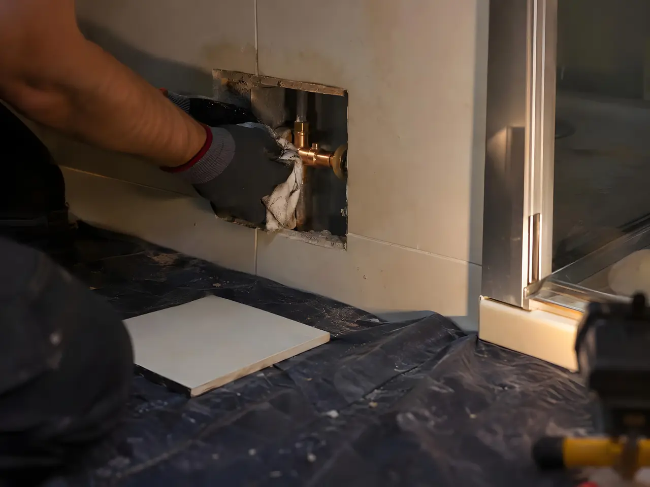 Main gantée qui essuie un raccord en laiton par une trappe ouverte dans la faïence, au pied d'une douche