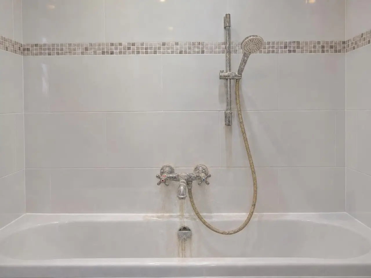 Vieux mélangeur de baignoire à deux têtes couvert de calcaire, flexible jauni, coulure sur l'émail