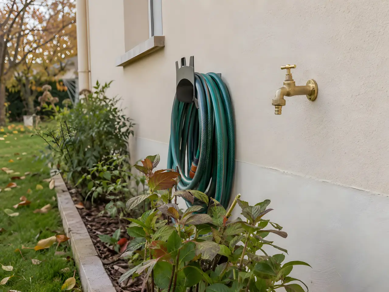Robinet extérieur en laiton neuf sur une façade enduite beige, tuyau d'arrosage vert enroulé sur son support