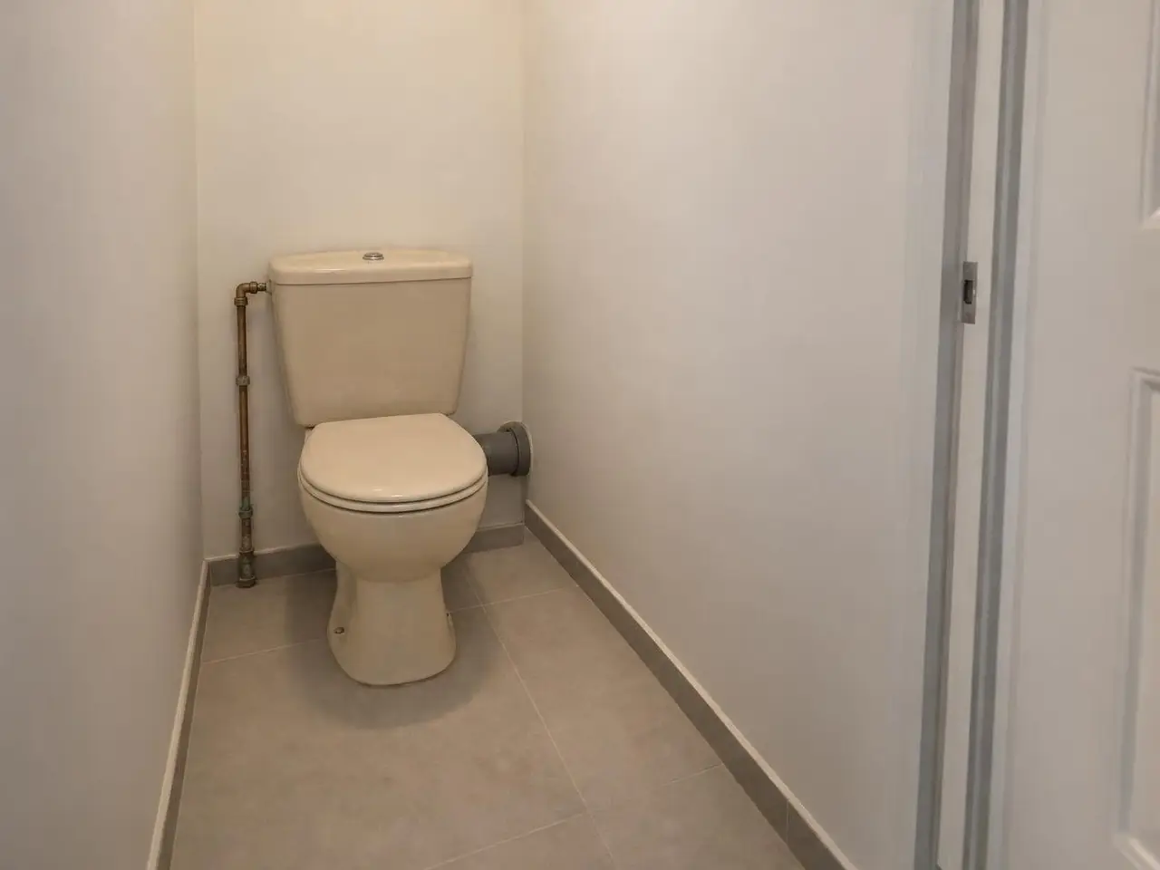 Petites toilettes aux murs blancs, vieux WC au sol beige à réservoir, tuyau de cuivre et pipe grise apparents